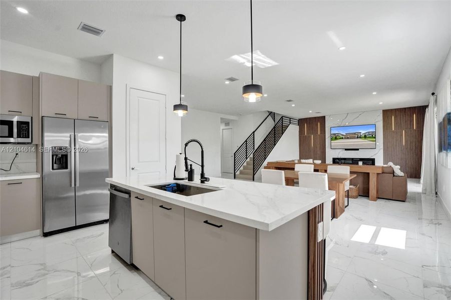 Open-concept living space with large format tile flooring, a kitchen island with stone countertop, stainless steel appliances, a modern staircase with metal railings, and a feature wall with integrated lighting