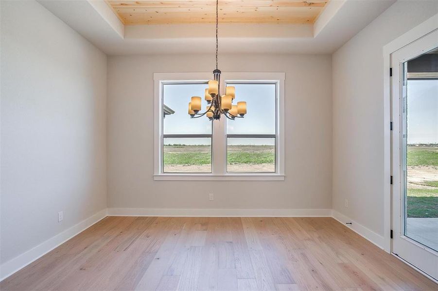 Unfurnished room with light wood-type flooring, a raised ceiling, an inviting chandelier, and baseboards Unfurnished room with light wood-type flooring, a raised ceiling, an inviting chandelier, and baseboards