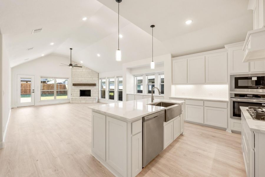 Open kitchen with white cabinets, large island, and stainless appliances open to living room with stone fireplace