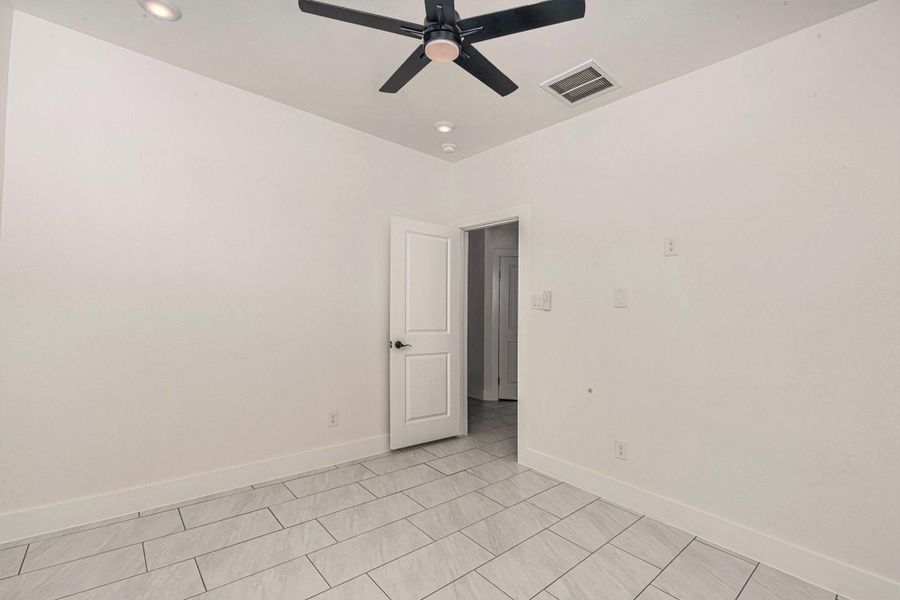 This is a bright, unfurnished room featuring light-colored tile flooring and white walls. It has a ceiling fan, recessed lighting, and a door leading to another area of the house.