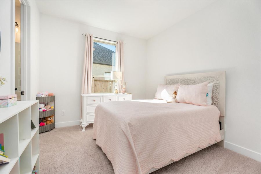 This cozy bedroom features a soft, neutral color palette with a comfortable bed, plush carpeting, and ample natural light from a window. It includes a dresser and storage shelves, perfect for a child's room or guest space.