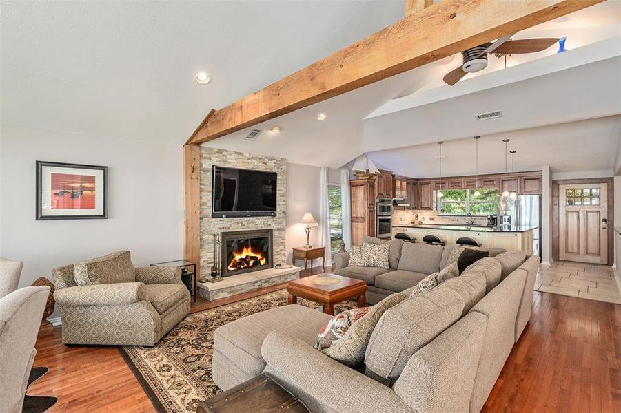 Living area featuring a stacked stone fireplace with hearth, wood-finish flooring, vaulted ceilings with exposed wood beams, and a ceiling fan