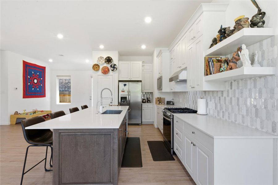 This kitchen features a modern design with sleek white cabinetry, a large island with a sink. It includes stainless steel appliances and a stylish tile backsplash. Open shelving adds character, while recessed lighting keeps the space bright.