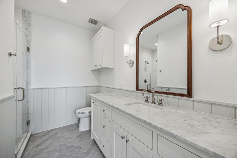 Timeless finishes define this secondary bath with a marble countertop vanity, polished nickel fixtures, and classic paneled wainscoting. Glass enclosed shower with marble trim and herringbone tile flooring adds subtle texture and a tailored touch.