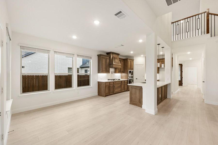Open-concept living area and kitchen with dark wood cabinets, light hardwood floors, and high ceilings with upper stair railing visible. Open-concept living area and kitchen with dark wood cabinets, light hardwood floors, and high ceilings with upper stair railing visible.