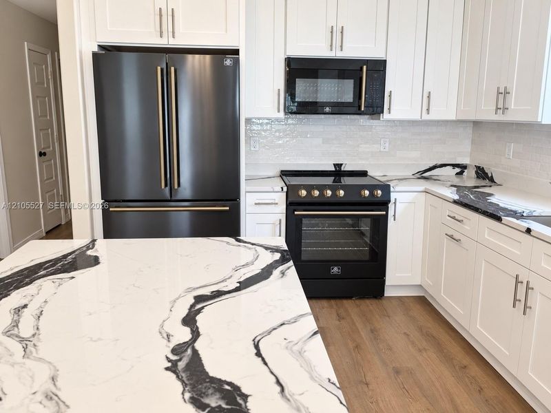 White shaker cabinetry with matte black appliances, wood-finish flooring, and a patterned countertop White shaker cabinetry with matte black appliances, wood-finish flooring, and a patterned countertop