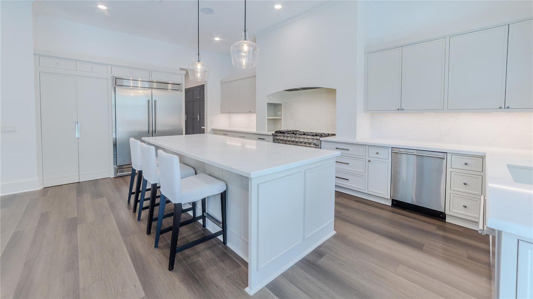 This modern kitchen features sleek white cabinetry, a spacious island with seating, stainless steel appliances, and elegant pendant lighting. The open layout and light wood flooring create a bright and inviting space.
