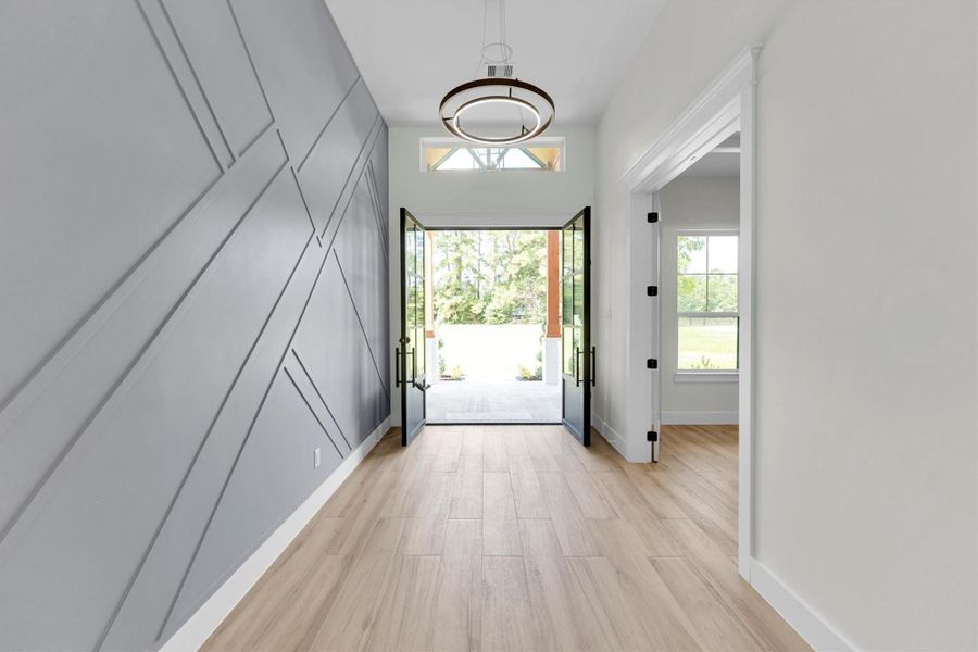 This entryway features a modern design with a stylish geometric accent wall, light wood flooring, and large glass double doors that open to a bright outdoor view, creating a welcoming and airy atmosphere.