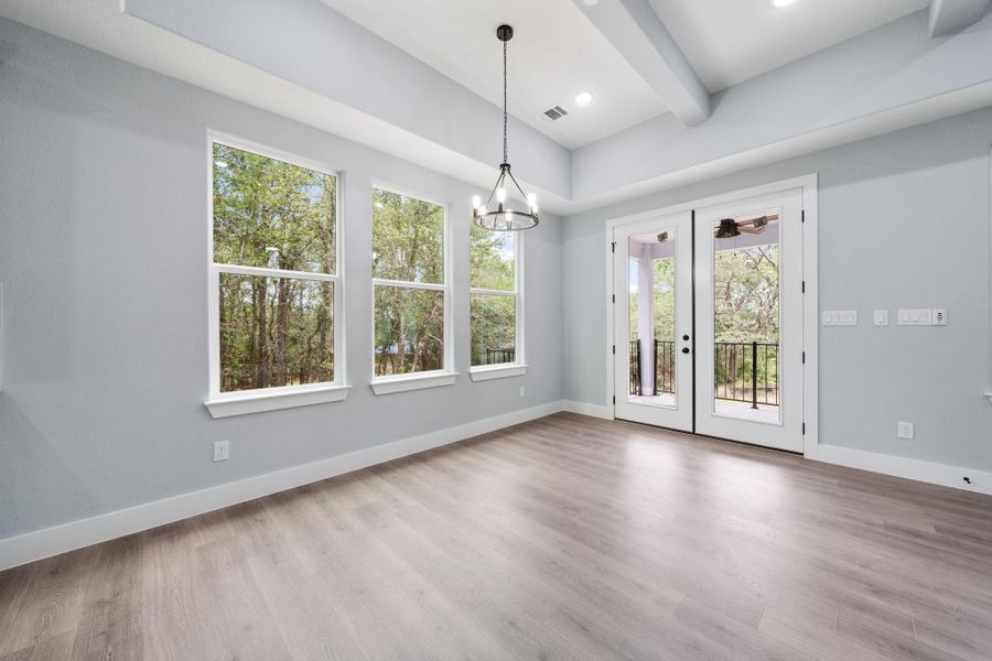 Interior space featuring wood-finish flooring, three large windows, double glass panel doors opening to an outdoor balcony with black railing, a vaulted ceiling with exposed beams, and a modern chandelier
