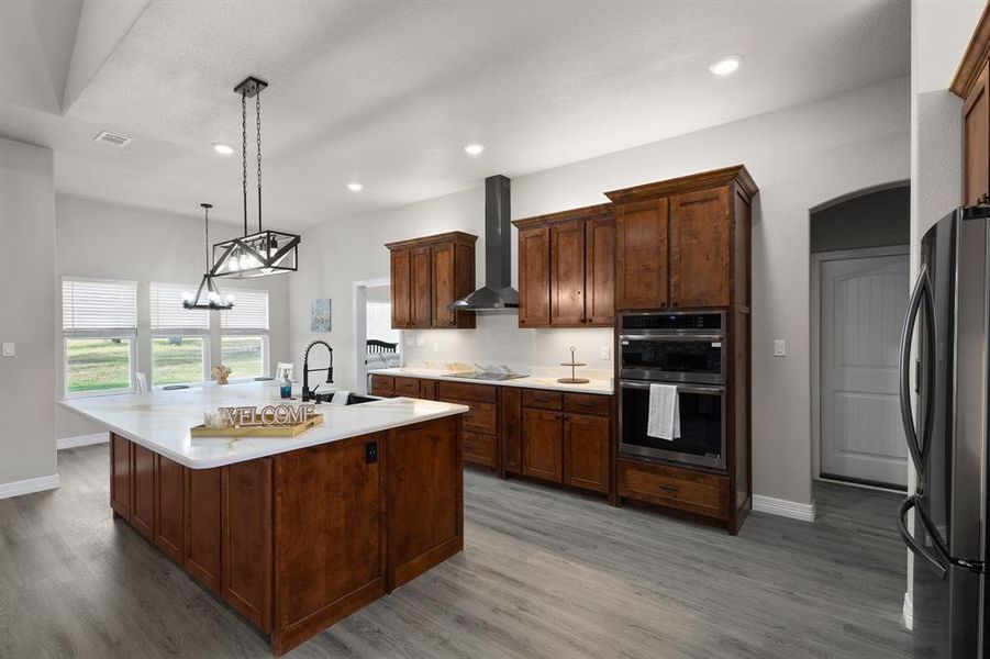 Kitchen with pendant lighting, appliances with stainless steel finishes, wall chimney exhaust hood, a kitchen island with sink, and a chandelier