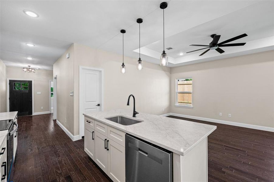 Open-concept kitchen and living area featuring dark wood-finish flooring, a kitchen island with a built-in sink and stainless steel dishwasher, pendant lighting, and a tray ceiling with a modern ceiling fan