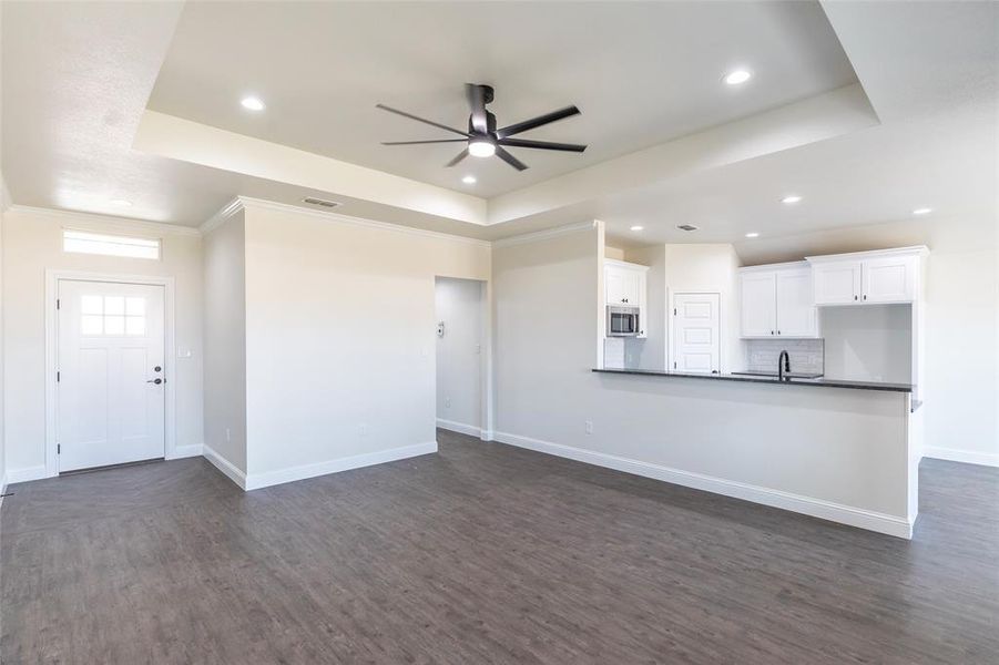 Open-concept interior featuring wood-finish flooring, a tray ceiling with recessed lighting and ceiling fan, and a kitchen bar with white cabinetry, a built-in microwave, and a dark countertop