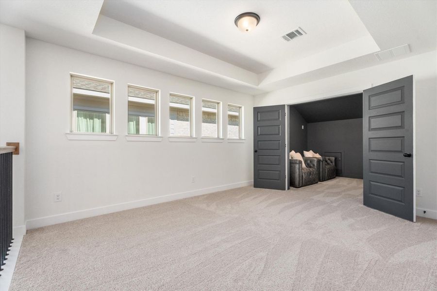 This spacious room features a neutral color scheme with plush carpeting, a row of upper windows for natural light, and a tray ceiling with recessed lighting. Double doors open to a cozy, dark-walled nook suitable for a home theater or reading area.