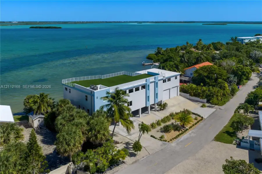Waterfront multi-story residence featuring a rooftop terrace with synthetic turf, perimeter railing, paved driveway, and attached two-car garage Waterfront multi-story residence featuring a rooftop terrace with synthetic turf, perimeter railing, paved driveway, and attached two-car garage
