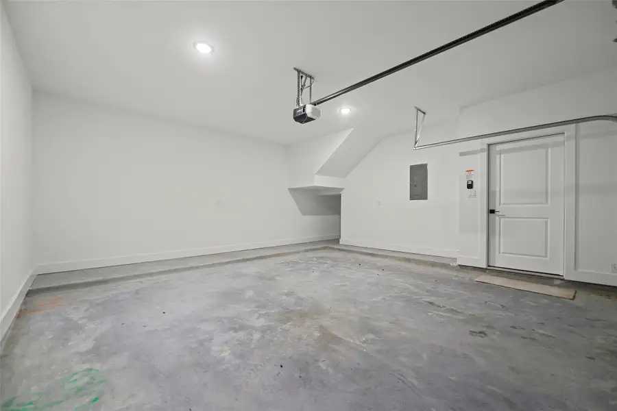 This photo shows a spacious, clean, and well-lit garage with concrete flooring, white walls, and a door leading inside. It features a garage door opener and ample space for parking or storage.