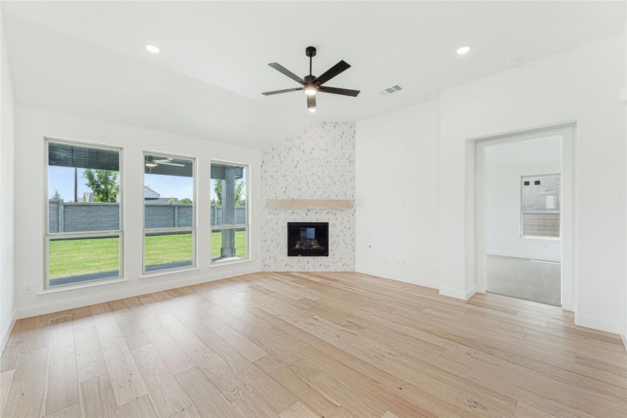Open living room with light hardwood floors, tile fireplace, ceiling fan, and large windows overlooking backyard Open living room with light hardwood floors, tile fireplace, ceiling fan, and large windows overlooking backyard