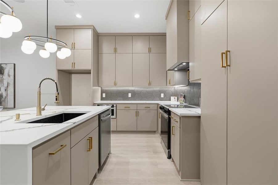 Open-concept kitchen featuring light-toned cabinetry with gold-finish hardware, a large island with a black sink and gold-finish faucet, white stone countertops, and a gray hexagonal tile backsplash
