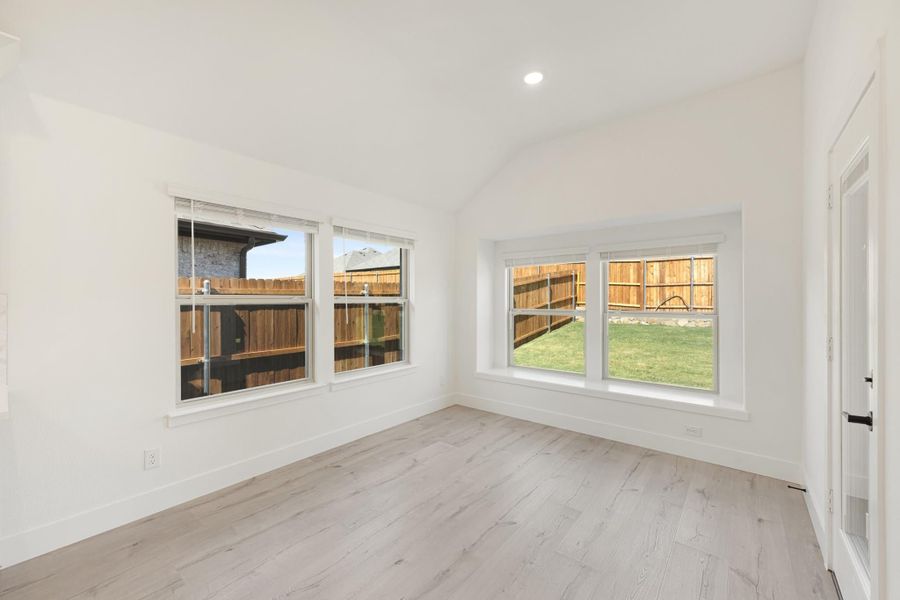 Empty room with vaulted ceiling, light wood floors, and large windows overlooking a fenced backyard