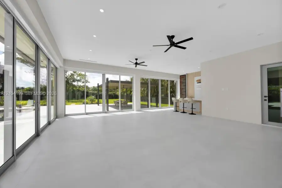 Large-format porcelain tile flooring, expansive floor-to-ceiling sliding glass doors, built-in wet bar with wood-panel detailing, recessed lighting, and dual matte black ceiling fans