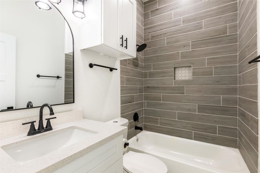 This bathroom features a modern design with sleek gray tile walls, a white vanity with a black faucet, and a matching black showerhead. The space includes a bathtub, built-in shelf, and stylish lighting, creating a clean and contemporary feel.