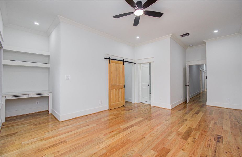 This spacious primary bedroom features beautiful hardwood floors, a ceiling fan, and a modern barn door. It includes built-in shelves and a desk area, making it ideal for a private home office or study.