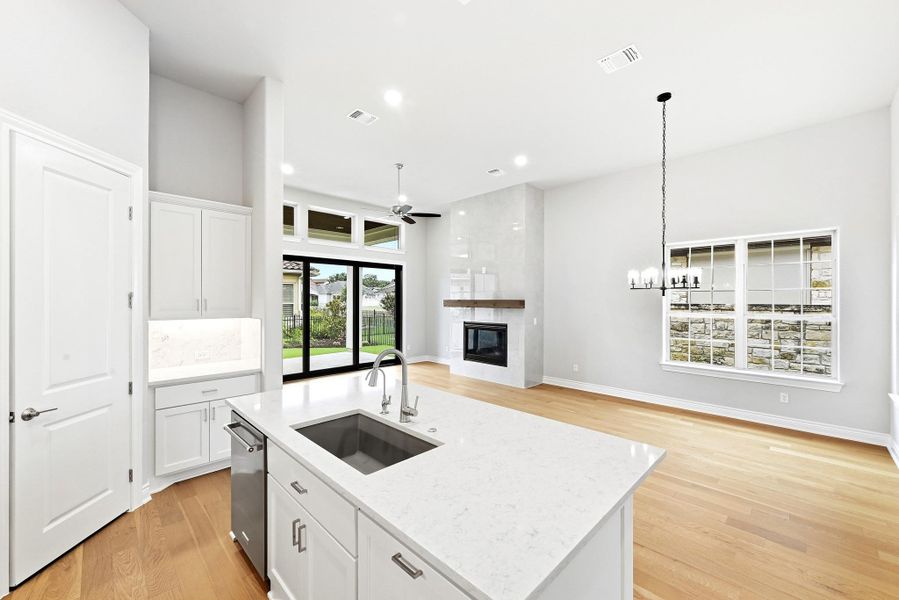 Open-concept living space featuring a kitchen island with an integrated sink, a built-in dishwasher, white cabinetry, wood-finish flooring, and a fireplace with a wood mantel
