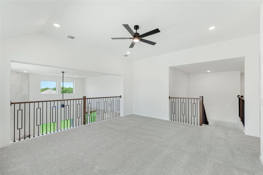 Open upstairs loft with gray carpet, ceiling fan, and black iron stair railing overlooking lower level Open upstairs loft with gray carpet, ceiling fan, and black iron stair railing overlooking lower level