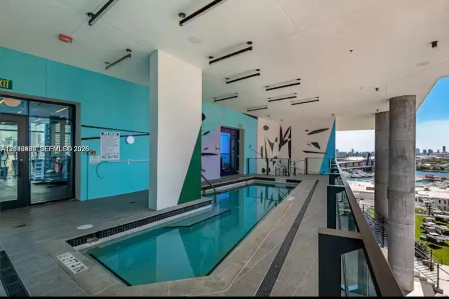 Covered outdoor pool featuring grey stone-finish tile decking, structural concrete support columns, a glass railing system, and linear recessed lighting Covered outdoor pool featuring grey stone-finish tile decking, structural concrete support columns, a glass railing system, and linear recessed lighting