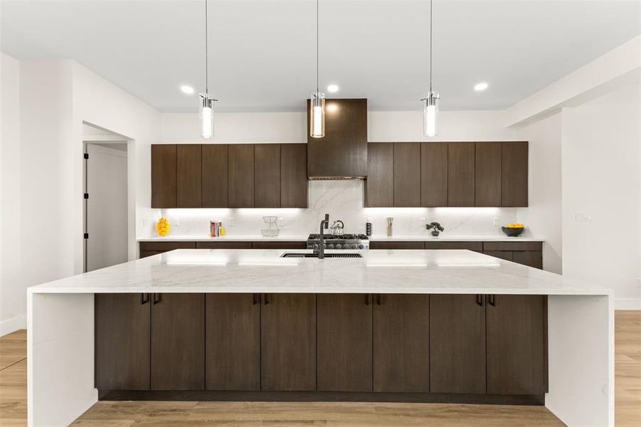 Kitchen featuring a large waterfall island with a light-toned countertop, dark wood-finish cabinetry, a gas range with an overhead vent hood, a light-toned backsplash, and recessed and pendant lighting