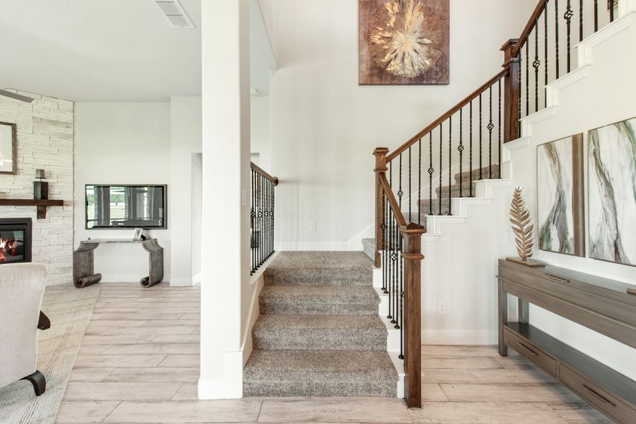 Carpeted staircase with wood and iron railing in open foyer with stone fireplace visible in background