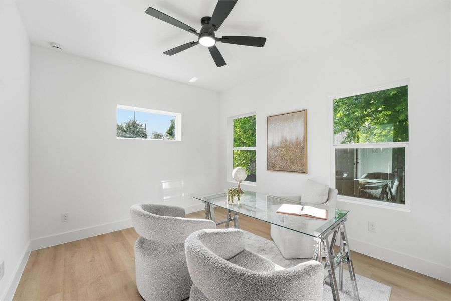 Bright and versatile home office or study featuring wood-look flooring, modern ceiling fan, clean white walls, and multiple windows that fill the space with natural light and serene tree views.