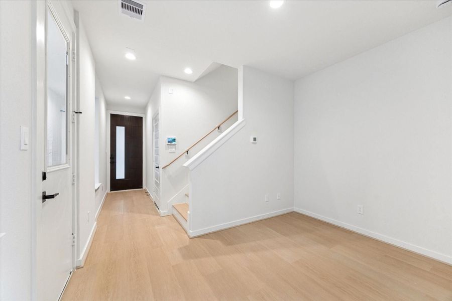 From the guest suite/flex space, the view toward the front door showcases the home’s clean, modern style. Light oak–style LVP floors and white trim create a bright, cohesive look, while the open staircase adds architectural interest. The sightline to the entry feels open and inviting, offering a spacious, connected feel as you look back toward the front of the home.