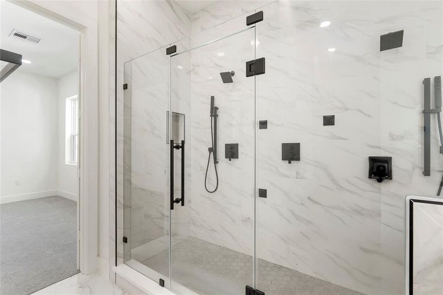 This is a modern bathroom featuring a spacious walk-in shower with frameless glass doors and marble-look tile walls. It has multiple shower heads and sleek black hardware. A small window provides natural light.