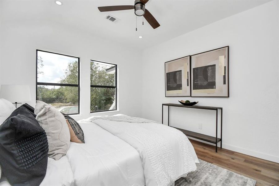 This is a bright and modern bedroom with clean lines, featuring a ceiling fan, large windows for natural light, and hardwood floors. It includes a cozy bed with stylish bedding and has direct access to what appears to be an en-suite bathroom.