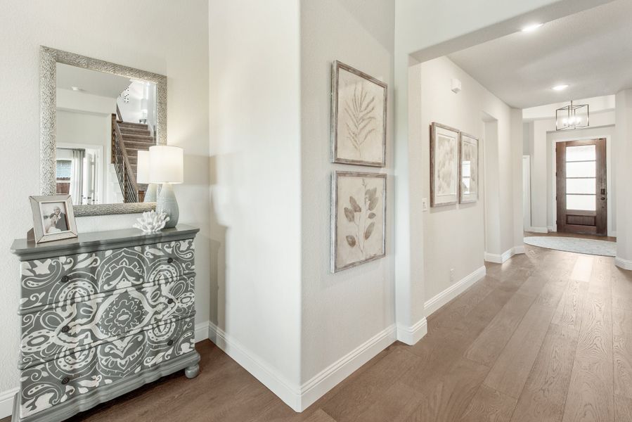 Bright home entryway with hardwood floors, decorative dresser, wall art, and wood front door at end of hallway