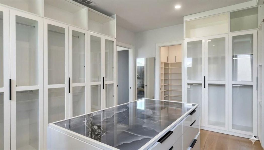 Custom closet system featuring a central island with a multi-toned countertop, extensive white cabinetry with glass-front doors, modern black hardware, recessed lighting, and light wood-finish flooring