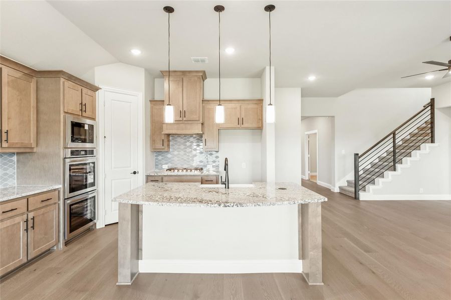 Kitchen with large granite island, light wood cabinets, stainless steel appliances, and hardwood floors open to staircase area Kitchen with large granite island, light wood cabinets, stainless steel appliances, and hardwood floors open to staircase area