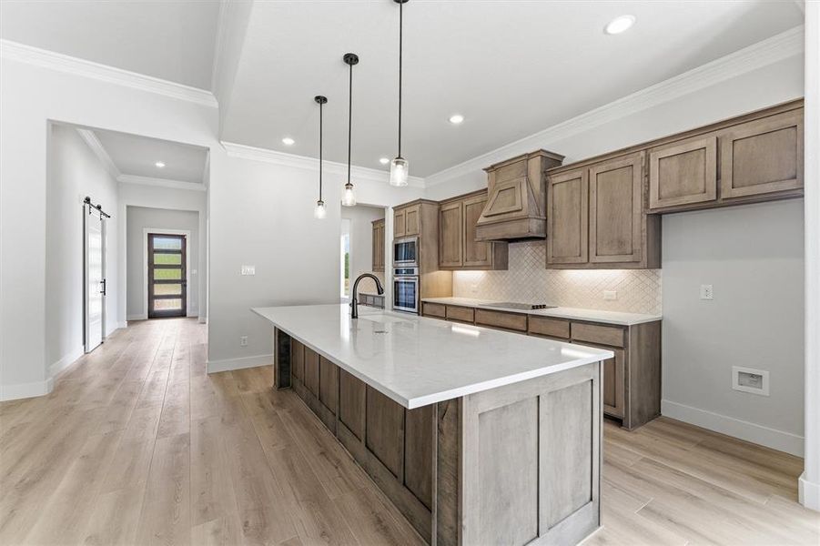 Kitchen featuring a center island with sink, wood finish cabinetry, a barn door, light wood-type flooring, and crown molding