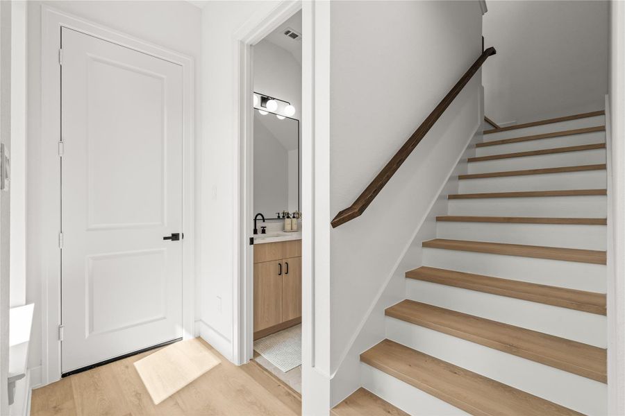 This view shows how the main floor connects. The light oak flooring leads past a white door toward a private, tucked-away powder room. To the right, a staircase with white risers, oak steps, and a matching wood handrail leads to the upper level.
