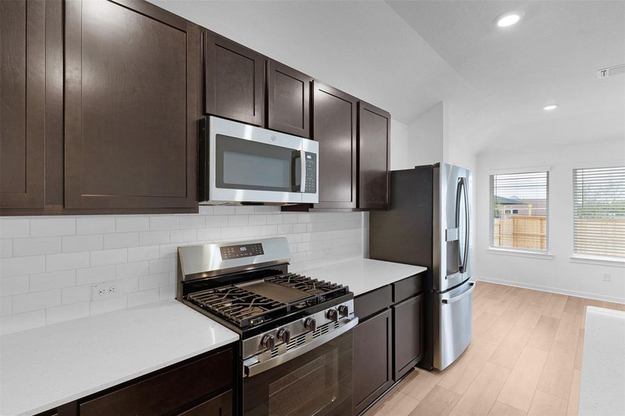 Kitchen with appliances with stainless steel finishes, dark brown cabinetry, lofted ceiling, decorative backsplash, and light wood-style floors Kitchen with appliances with stainless steel finishes, dark brown cabinetry, lofted ceiling, decorative backsplash, and light wood-style floors