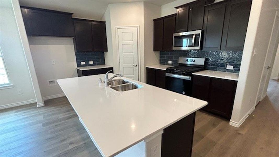 Kitchen featuring a large central island with an undermount double-basin sink, dark wood-finish cabinetry, solid surface countertops, a mosaic tile backsplash, and wood-finish flooring