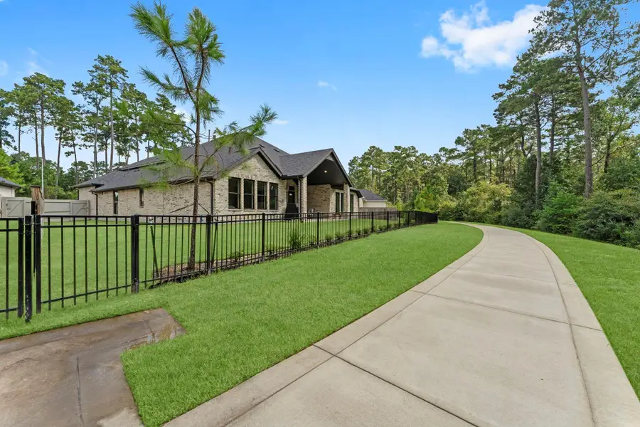 Just beyond the backyard fence is a long, winding concrete path stretching through a lush green lawn. A black metal fence runs along the path on the left, with a small tree and some landscaping. The background is filled with a dense line of tall pine trees, giving the yard a private, park-like feel. Just beyond the backyard fence is a long, winding concrete path stretching through a lush green lawn. A black metal fence runs along the path on the left, with a small tree and some landscaping. The background is filled with a dense line of tall pine trees, giving the yard a private, park-like feel.