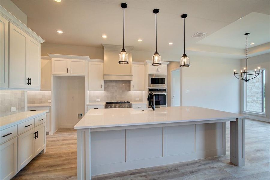 Kitchen with light hardwood / wood-style floors, appliances with stainless steel finishes, decorative light fixtures, a large island with sink, and tasteful backsplash