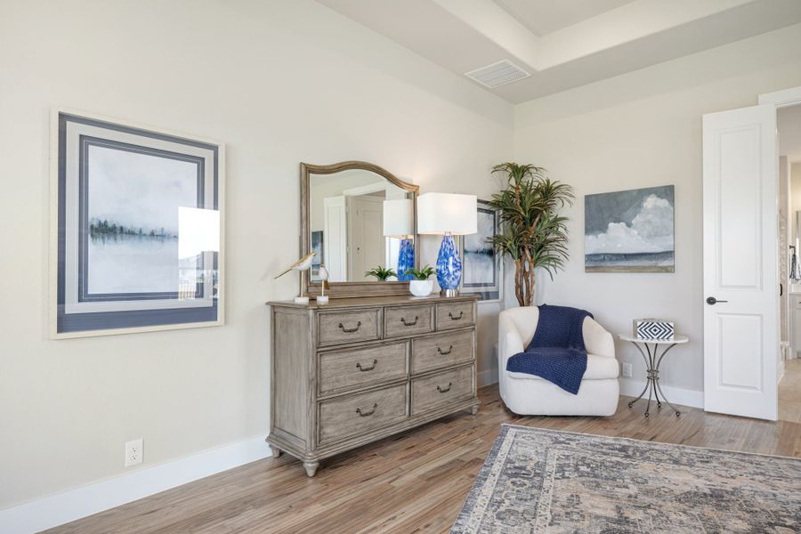 Bedroom sitting area with wood dresser, mirror, white accent chair, and hardwood floors under a tray ceiling