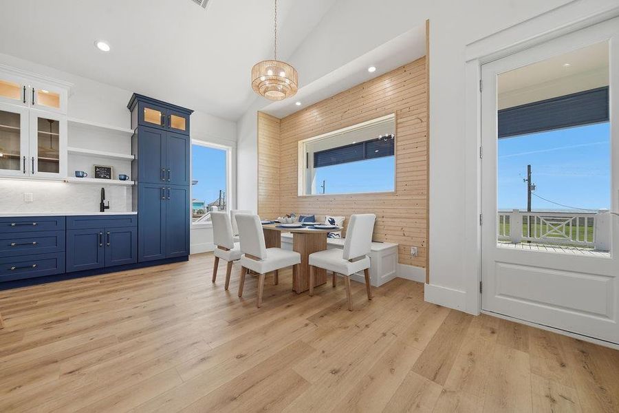 This bright dining area features a cozy built-in wooden nook, modern white and blue cabinetry, and ample natural light from large windows and a glass door leading to an outdoor space. The light wood flooring and stylish pendant light create a warm, inviting atmosphere.