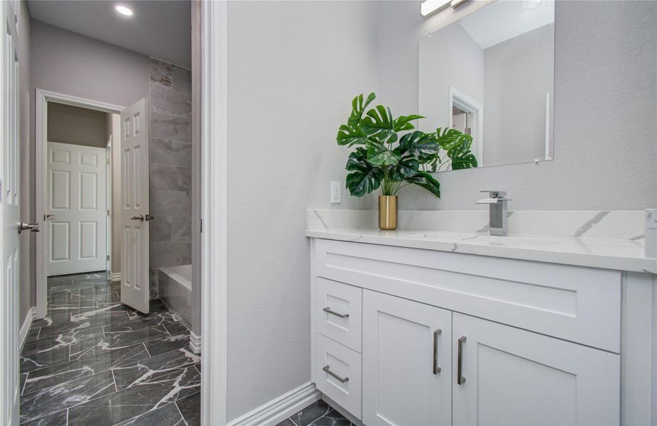 This bathroom features a sleek design with marble-patterned floors, a modern white vanity with dual sinks, and a large mirror. The space is accented by a potted plant and has access to a bathtub area with tiled walls.
