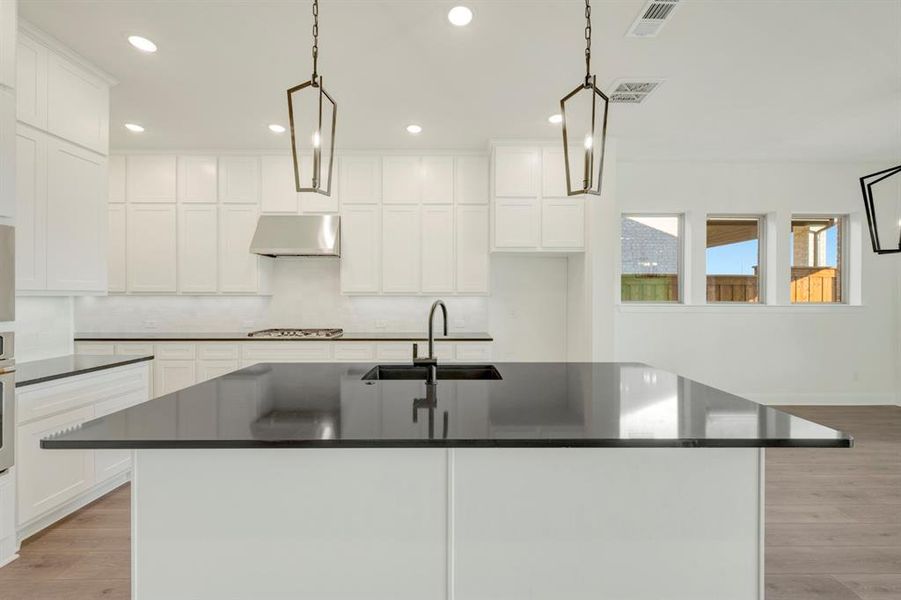 Kitchen featuring light wood finished floors, white cabinets, a center island with sink, and hanging light fixtures Kitchen featuring light wood finished floors, white cabinets, a center island with sink, and hanging light fixtures