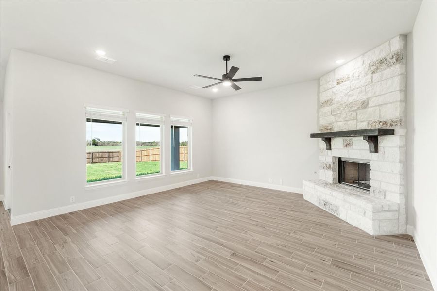 Open living room with wood-look floors, stone fireplace, ceiling fan, and large windows overlooking a fenced backyard. Open living room with wood-look floors, stone fireplace, ceiling fan, and large windows overlooking a fenced backyard.