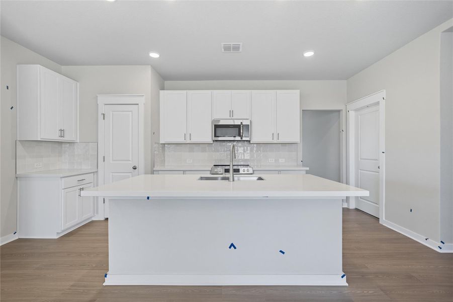 Kitchen with white cabinets, a kitchen island with sink, dark wood-type flooring, backsplash, and stainless steel microwave Kitchen with white cabinets, a kitchen island with sink, dark wood-type flooring, backsplash, and stainless steel microwave