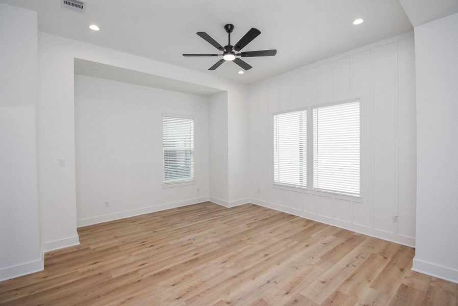 Bright, spacious room with light wood flooring, white walls, and a modern ceiling fan. Features large windows with blinds, recessed lighting, and a cozy alcove.